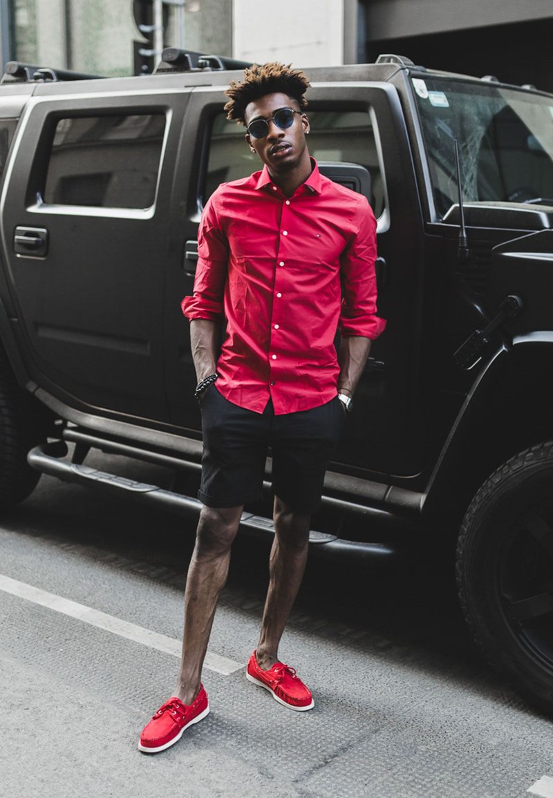 A man in a red button-up shirt and black shorts stands beside a black SUV. He is wearing round sunglasses and red shoes with white soles.