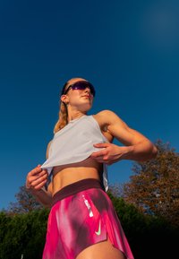 Sportovní oblečení s světle šedým topem a šortkami s růžovým gradientem. Šortky mají tmavý pás a zelené logo Nike.