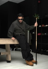 Schwarze Jeansjacke mit zwei vorderen Taschen, weite schwarze Cargo-Hosen und braune Stiefel. Mit einem weißen Stock und in einer minimalistischen Innenumgebung gestaltet.