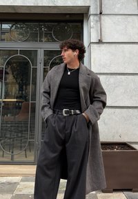 Mann iført grå lang frakk, svart skjorte, vide grå bukser og AirPods, står med hendene i lommene nær glassdør og plantekasse.