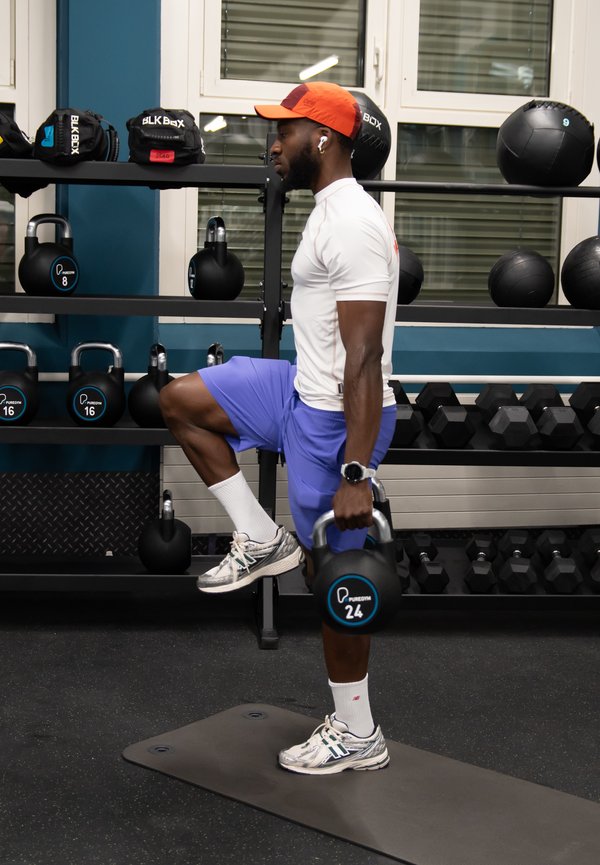 Homme portant une casquette orange relève le genou gauche tout en tenant un kettlebell de 24 kg dans une salle de sport avec des kettlebells et des ballons médicinaux sur des étagères.