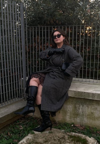 Abrigo de punto en gris con cinturón, falda de encaje debajo, botas de cuero negras hasta la rodilla y guantes negros. Incluye gafas de sol y un bolso pequeño.