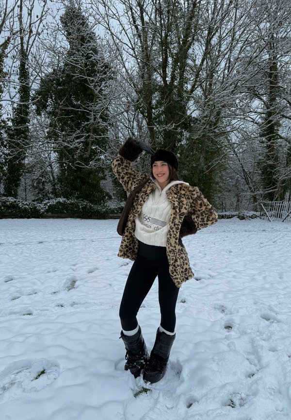 Kobieta w płaszczu w panterkę, białym swetrze, czarnych legginsach i zimowych butach stoi na śniegu, uśmiechając się, na tle pokrytych śniegiem drzew.