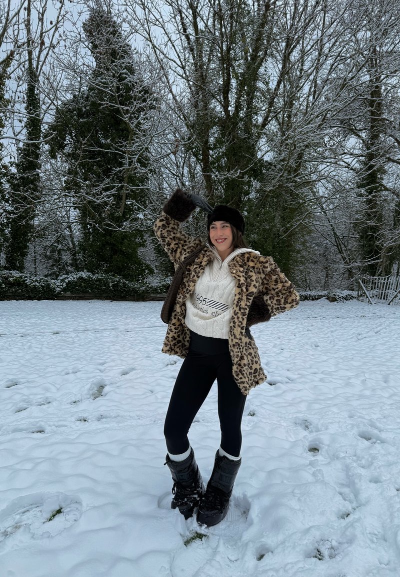 Femme portant un manteau imprimé léopard, un pull blanc, des leggings noirs et des bottes d'hiver, debout sur la neige, souriante, avec des arbres couverts de neige en arrière-plan.