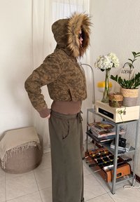 Personne portant une veste à capuche camouflage avec bordure en fourrure ajustant son pantalon, debout à côté d'une étagère métallique avec des plantes, des magazines et une table de baby-foot.