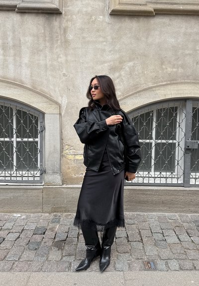 Mujer con chaqueta negra de cuero, falda midi, botas y pequeñas gafas de sol, de pie en una calle adoquinada junto a ventanas con rejas.