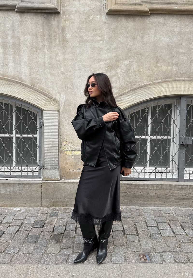 Woman wearing black leather jacket, midi skirt, boots, and small sunglasses stands on cobblestone street by barred windows.