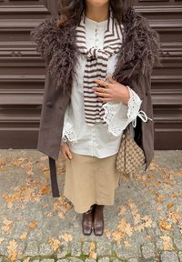 Brun, strukturert kåpe med pelskrage, hvit bluse med blonder på mansjettene, stripete skjerf, beige skjørt, brune knelange støvler og mønstret veske.