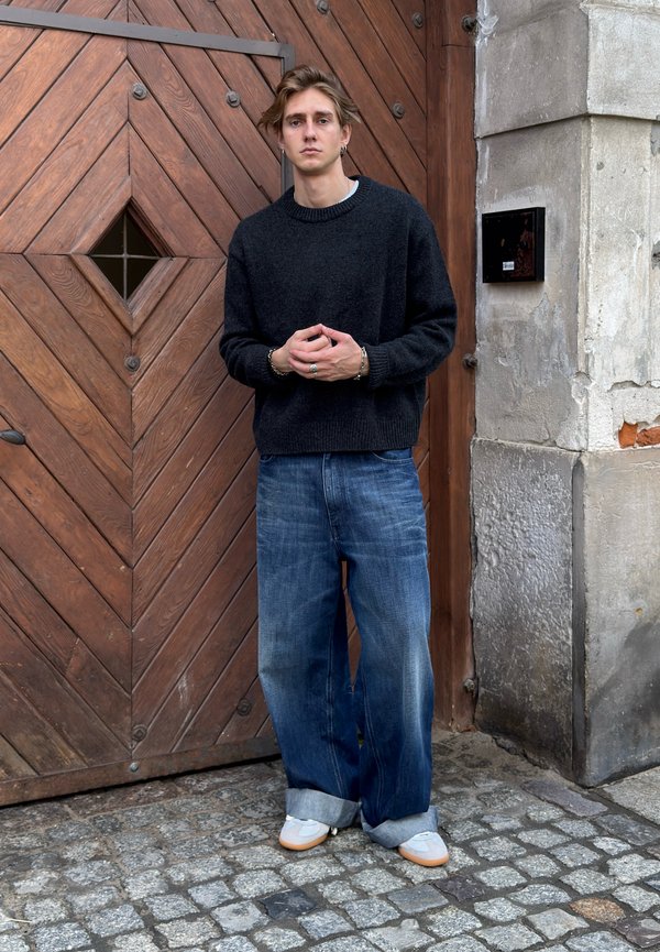 Jeune homme aux cheveux mi-longs se tient devant une grande porte en bois, portant un pull noir, un jean large bleu et des baskets blanches.