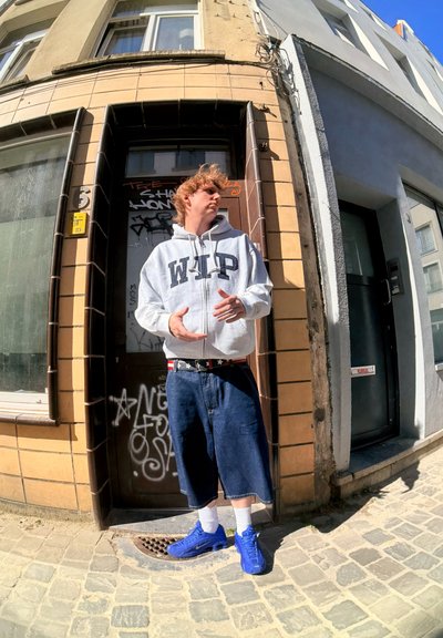 Sudadera gris con cremallera y texto "WIP", pantalones cortos de mezclilla azul, zapatillas deportivas azules y calcetines blancos. De pie frente a un edificio de azulejos con graffiti.