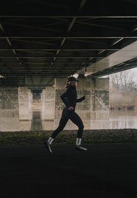Vrouw die jogt onder een betonnen brug bij een rivier, gekleed in zwarte sportkleding, met pet en witte sokken in een stedelijke omgeving met weinig licht.