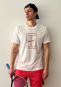 Camiseta de algodão branca com um logótipo vermelho da Nike em um contorno retangular. Combinada com calç�ões cor-de-rosa brilhantes. Segurando uma raquete de ténis.