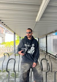 Felpa nera con stampa grafica bianca, pantaloni shorts in denim larghi e occhiali da sole. Tiene un piccolo sacchetto nero in un'area coperta.