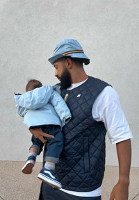 A man in a navy quilted vest holds a child in a light blue puffer jacket, both wearing casual shoes and a blue bucket hat.
