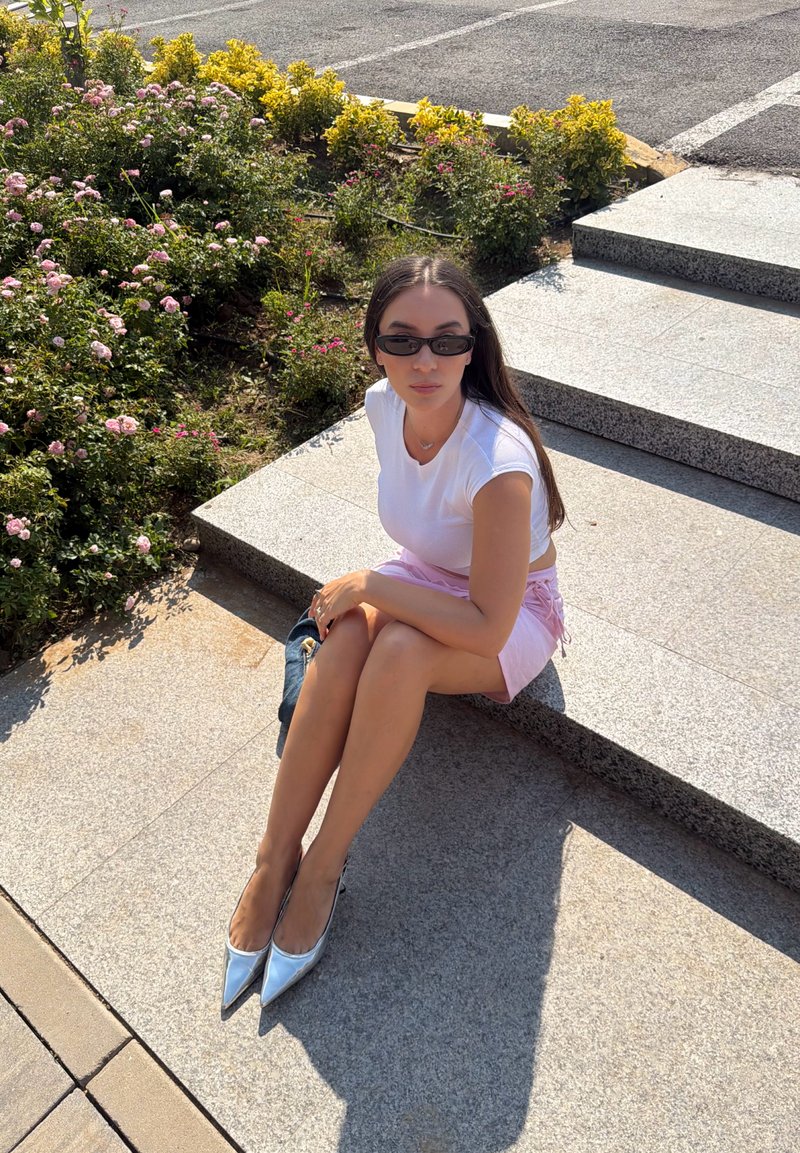 Jeune femme portant des lunettes de soleil, un haut blanc, une jupe rose et des talons argentés assise sur des marches en pierre en plein air à côté de buissons fleuris sous le soleil.