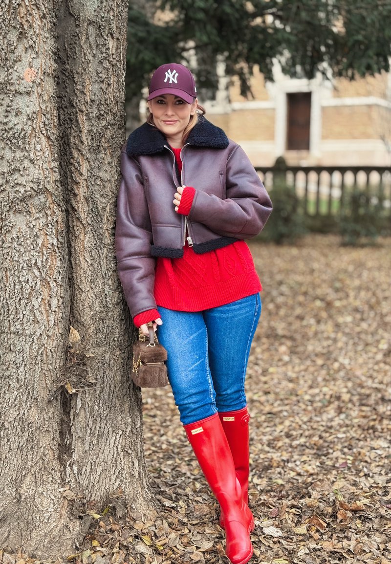 Braune Lederjacke, kurz geschnitten, mit schwarzem Futter, roter Strickpullover mit Zopfmuster, blaue Jeans und leuchtend rote knielange Gummistiefel; hält ein braunes Pelzaccessoire.