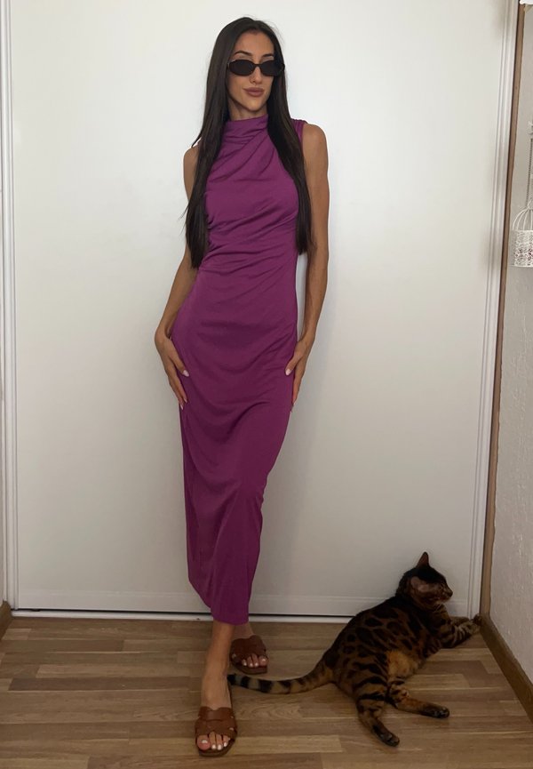 Femme en robe violette sans manches et sandales marron pose à l'intérieur à côté d'un chat tacheté marron et noir couché sur un sol en bois.