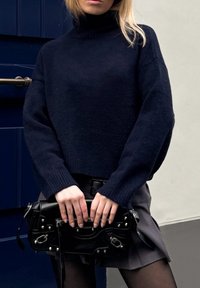 Tengerés turtleneck pulóver, esett vállakkal, textúrált kötésben, fekete kézitáskával, ezüst hardverekkel és cipzárokkal párosítva; szürke szoknya látható.