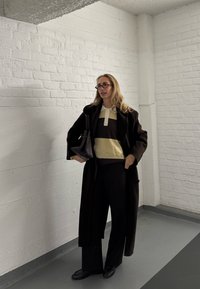 Hnedý oversize kabát, svetlohnedý a tmavohnedý pruhovaný sveter s bielym golierom, čierne nohavice a kabelka; nosený s okuliarmi; pozadie s jednoduchou bielou stenou.