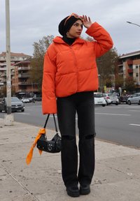 Veste en duvet orange, jeans noirs déchirés, sac à main noir avec écharpe orange, chaussures noires. Les accessoires comprennent des lunettes de soleil orange et des boucles d'oreilles.