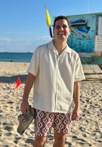 Χαμογελαστός άνδρας που κρατά σανδάλια σε ηλιόλουστη παραλία, φορώντας κοντομάνικο πουκάμισο με υφή και σορτς με λουλουδάτο μοτίβο, με τον ωκεανό και έναν πύργο ναυαγοσώστη στο βάθος.