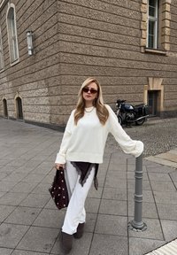 Bijeli pleteni džemper širokih rukava, upareni s višeslojnim bijelim hlačama i tamnocrvenom torbom ukrašenom velikim biserima; sivi čizme dovršavaju izgled.