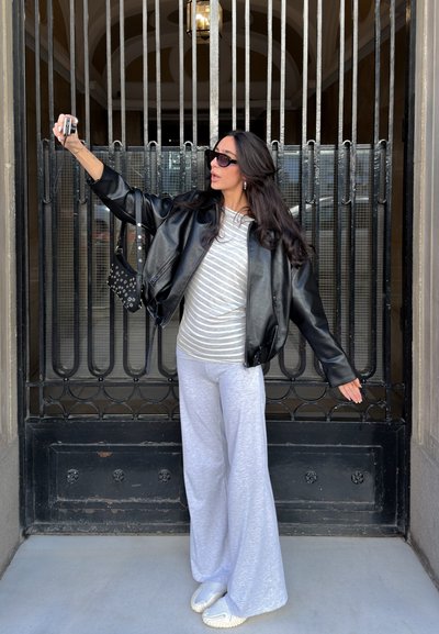 Mujer con gafas de sol, chaqueta de cuero negra, camiseta a rayas y pantalones anchos grises tomando una selfie con una cámara frente a una reja metálica.