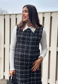 Haljina s crno-bijelim kariranim uzorkom, ispod bijela košulja s ovratnikom. Tkanina s teksturom, pripijeni kroj i zlatni gumbi. Crna torbica.