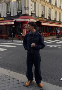 Mörkblå denimjacka med matchande löst sittande jeans, med stora fickor. Bruna arbetsskor med kontrasterande snören. Urban gatumiljö.