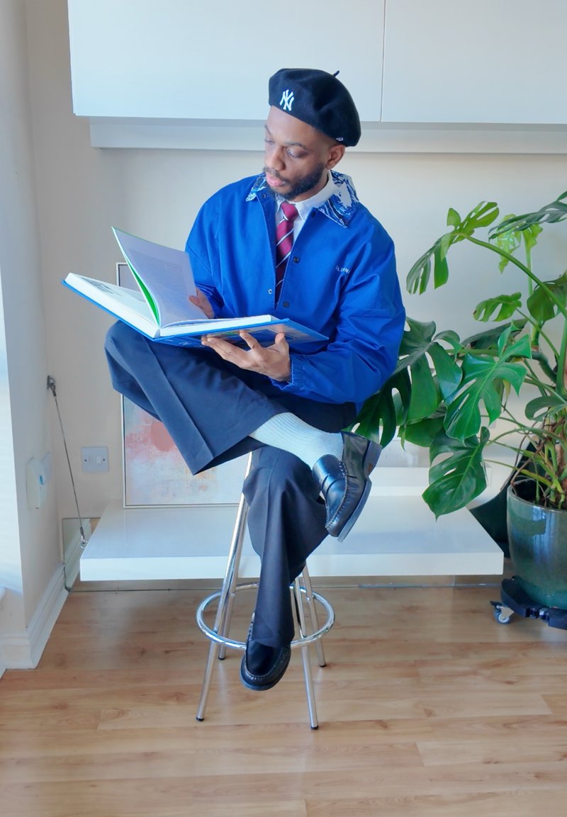 Homme assis en tailleur sur un tabouret lisant un grand livre ouvert, portant une veste bleue et un béret noir, à côté d'une grande plante verte.