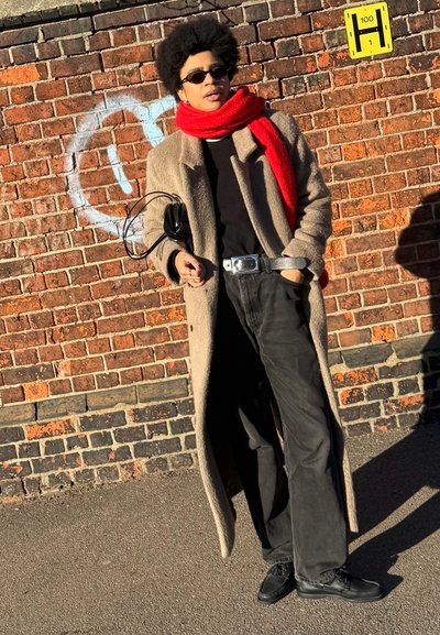 Persona con cabello rizado negro que lleva gafas de sol, bufanda roja, abrigo beige, conjunto negro, sosteniendo una pequeña bolsa negra, de pie frente a una pared de ladrillos.