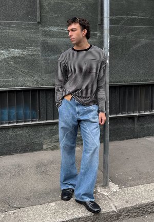 Hombre con camisa de rayas en blanco y negro de manga larga, jeans azules sueltos con un parche, zapatos negros, de pie en la acera junto a un poste de metal.