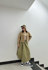 Μπεζ κοντό σακάκι με σκούρο γιακά, ανοιχτό μπεζ τοπ, ελκυστική πράσινη μακριά φούστα, μαύρα και λευκά αθλητικά παπούτσια και ελκυστικό καπέλο. Μαύρα γυαλιά ηλίου.