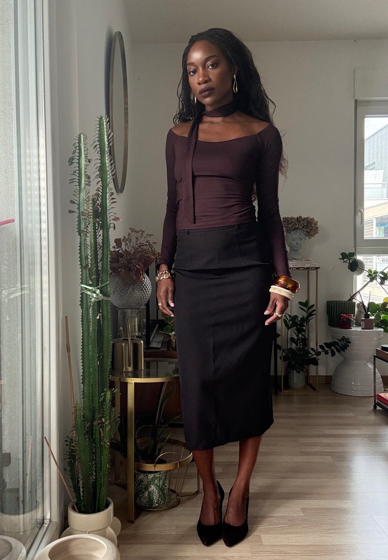 Langärmliges burgunderrotes Oberteil mit drapiertem Halstuch, kombiniert mit einem figurbetonten schwarzen Bleistiftrock und schwarzen High-Heels.