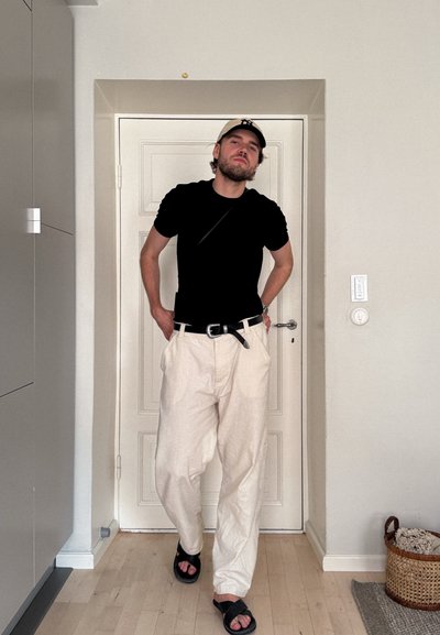 Camisa negra de manga corta, pantalones beige de corte holgado, cinturón negro, sandalias negras, de pie en una habitación de color claro junto a una puerta.