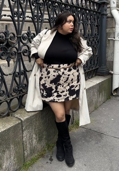 Mini falda de estampado de vaca beige con un cinturón negro, combinada con un suéter negro de cuello alto y un abrigo ligera beige; lleva botines negros hasta la rodilla.