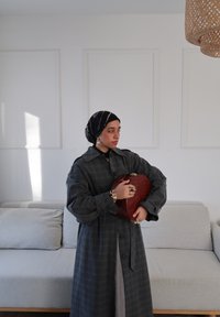 Long manteau gris à carreaux avec fermeture à boutons, porté sur un fond sombre, tenant un sac à main rouge texturé. Foulard noir et blanc à motifs.