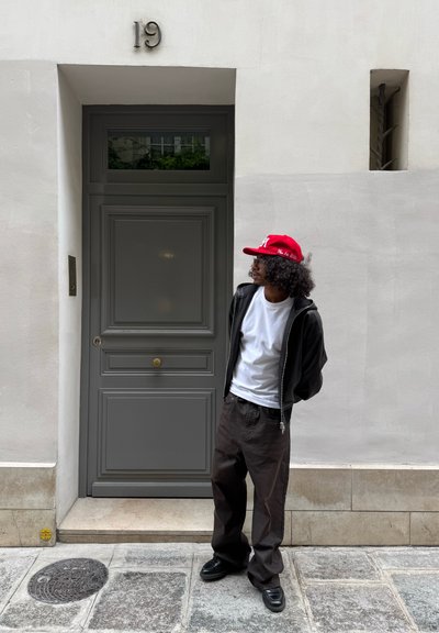 Puerta gris con un tirador en forma de sol, pared blanca, persona con una chaqueta negra y camisa blanca, gorra roja, pantalones marrones y zapatos negros.
