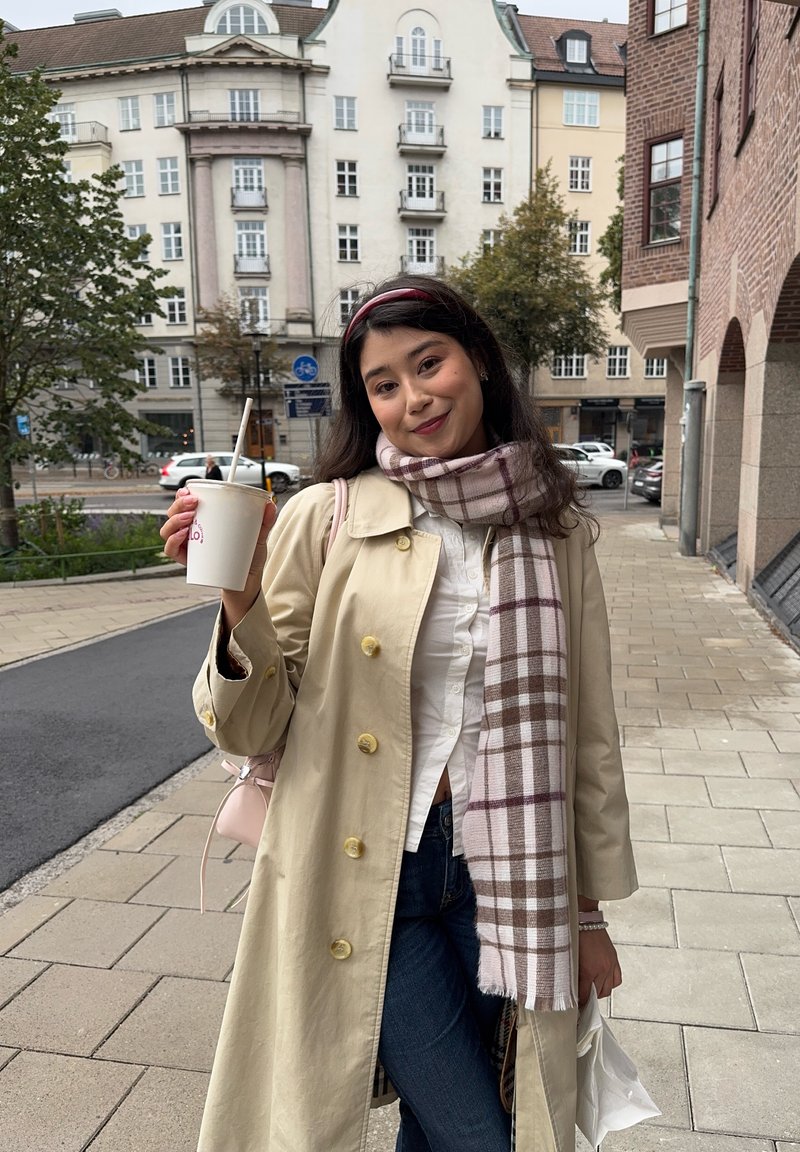 Beige Trenchcoat mit gelben Knöpfen, weißem Hemd und kariertem Schal. Hält eine Tasse mit einem Strohhalm. Der Hintergrund zeigt Ziegelmauer und moderne Gebäude.