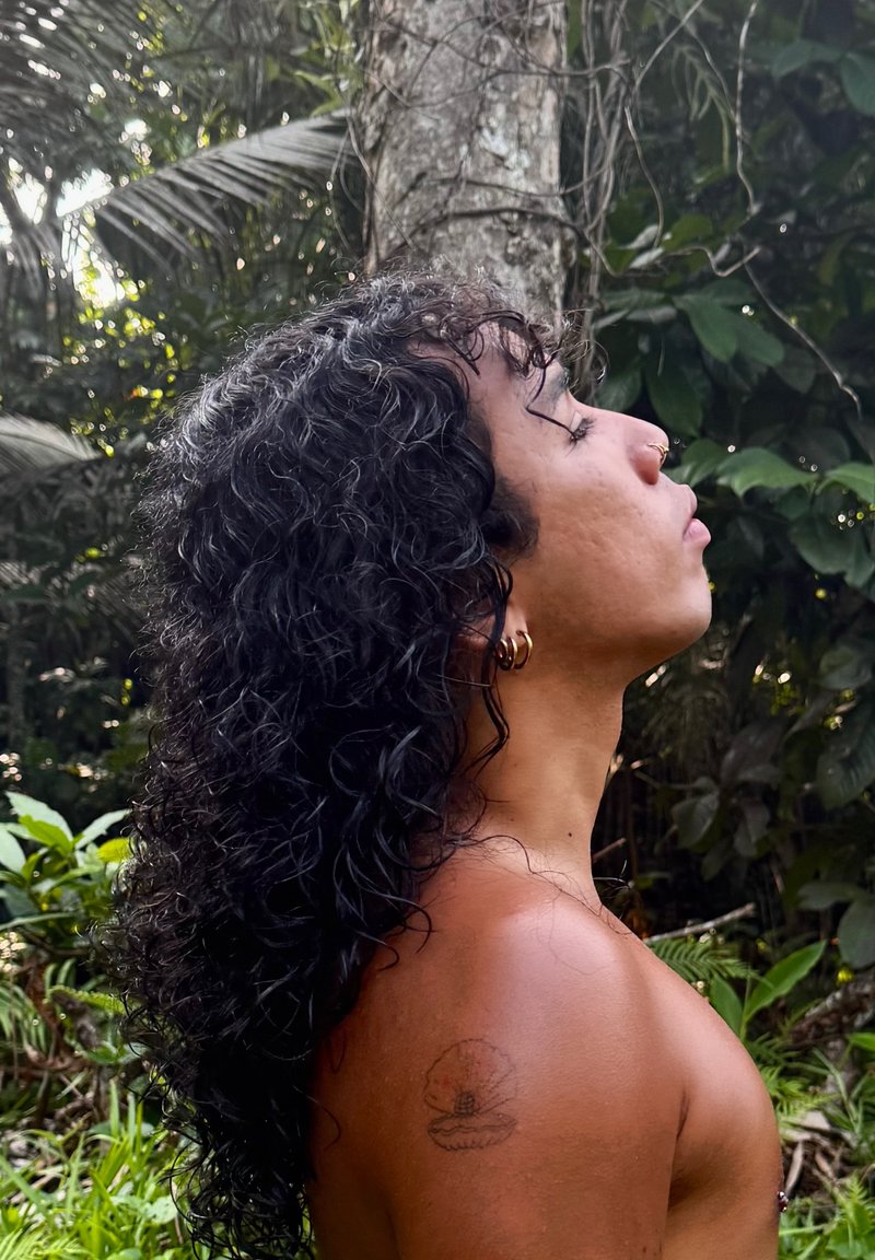 Persona con capelli lunghi e ricci, anello al naso e tatuaggio di conchiglia sulla spalla, in piedi in una foresta, occhi chiusi, rivolto verso destra.