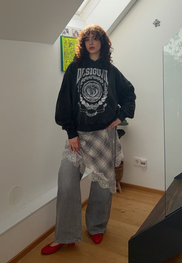 Krøllete dame står innendørs iført svart Desigual-hettegenser, rutete skjørt med blonder, løse grå bukser og røde sko nær trapp.