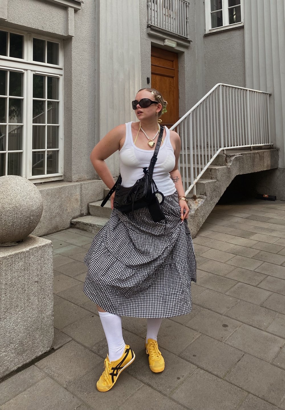 Svartvitt rutig kjol, vit linne, gula sneakers med svarta detaljer, svart crossbody-väska, iögonfallande solglasögon, flätad frisyr.