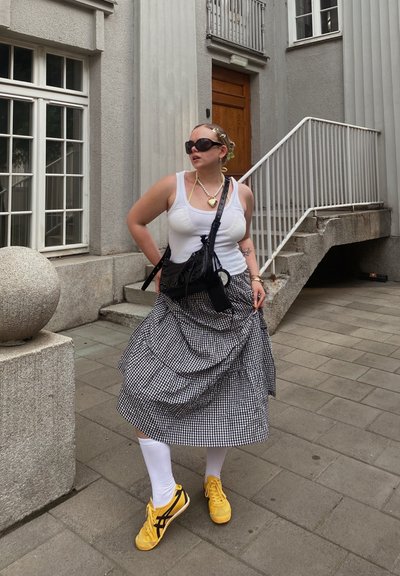 Falda de cuadros blanco y negro, camiseta sin mangas blanca, zapatillas amarillas con acentos negros, bolso negro de bandolera, gafas de sol llamativas, peinado trenzado.