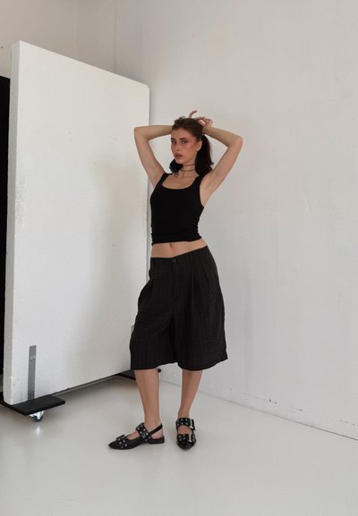 Top negro de tirantes combinado con pantalones cortos amplios y plisados en negro. El calzado presenta un diseño sin talón con detalles metálicos. Fondo blanco.