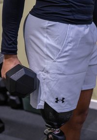 Light grey athletic shorts with side pockets, textured fabric, and black Under Armour logo. A person holds a black hex dumbbell.
