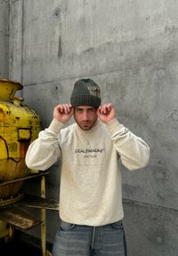 Light gray sweatshirt with printing, loose fit. Dark green ribbed beanie with a patch. Concrete background with a yellow industrial object.