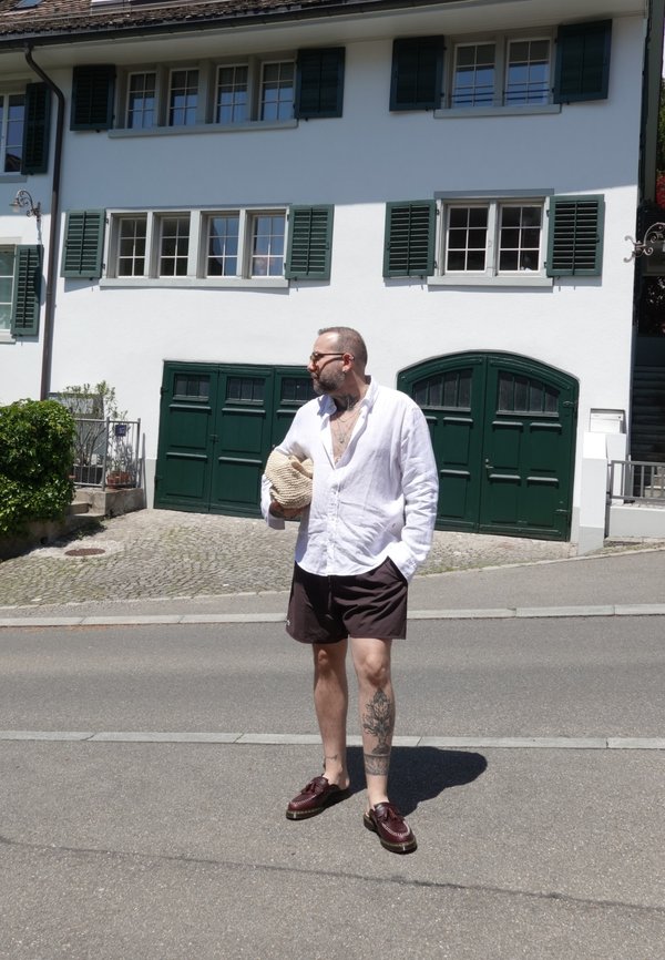 Mand med tatoveringer iført hvid skjorte og mørke shorts, der holder en hat, stående på gaden foran et hvidt hus med grønne skodder og garagedøre.