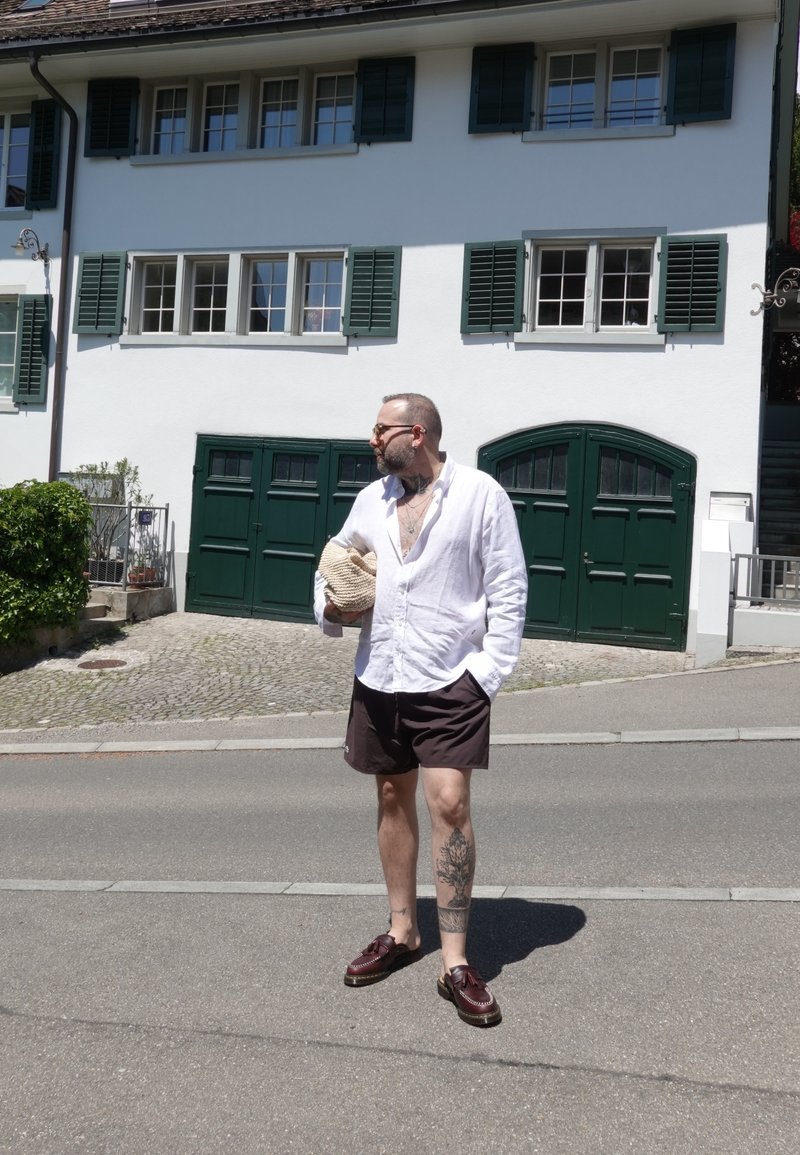 Homme avec des tatouages portant une chemise blanche et un short sombre, tenant un chapeau, debout dans la rue devant une maison blanche avec des volets verts et des portes de garage.