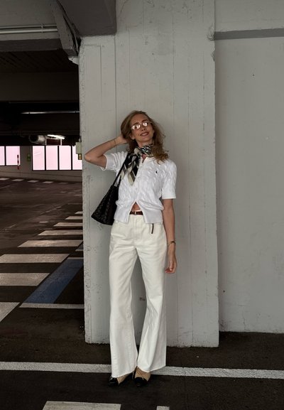 Jersey corto de punto trenzado blanco, pantalones amplios blancos, bufanda negra y blanca con estampado, bolso negro, gafas de sol. Fondo de garaje.