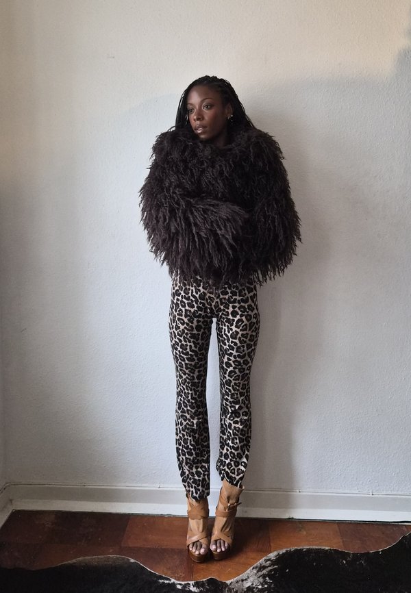 Une femme se tient devant un mur uni, vêtue d’un haut noir volumineux et hirsute, d’un pantalon à imprimé léopard et de sandales beiges à talons hauts.